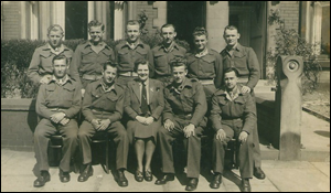 A group of ten men in military uniforms and one woman in civilian clothing sit and stand in two rows outdoors in front of a building.