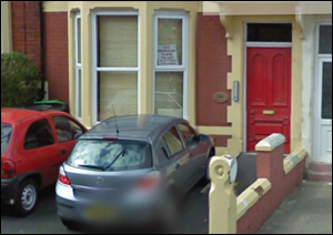 A grey car is parked in a small drive in front of a house with a red door; a red car is parked on the street beside it.