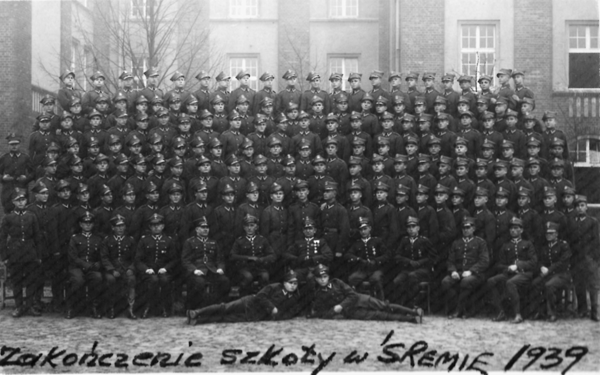 Black-and-white photo of a large group of uniformed men posing in rows outside a building; handwritten text at the bottom reads “Zakończenie szkoły w Śremie 1939.”.
