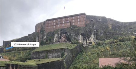 A large stone fortress sits atop a grassy hill, with a memorial plaque labelled "2DSP Memorial Plaque" visible on the lower left side.