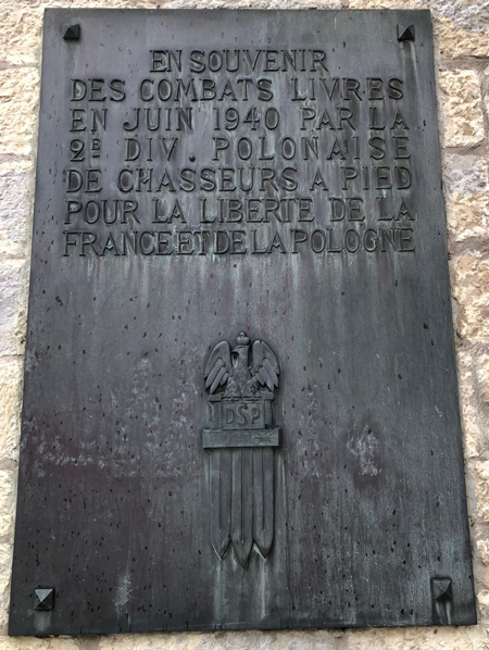 A metal plaque on a stone wall commemorates the 2nd Polish Infantry Division's battles in June 1940 for the freedom of France and Poland, featuring an eagle emblem and inscription in French.