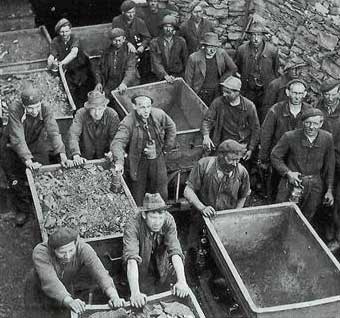 A group of workers in caps and overalls stand outdoors, pushing large metal trolleys filled with rocks or rubble, next to a stone wall.