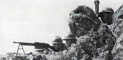 Four soldiers wearing helmets take cover behind rocks, with one aiming a machine gun over the rocks and the others observing.