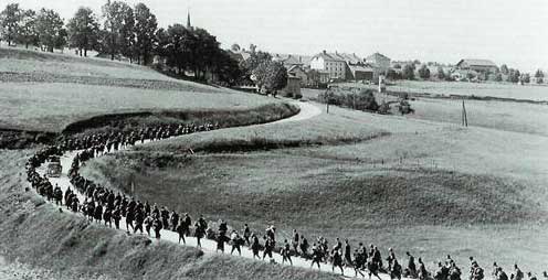 A large group of soldiers marches in a winding line along a rural road with fields and a village in the background.