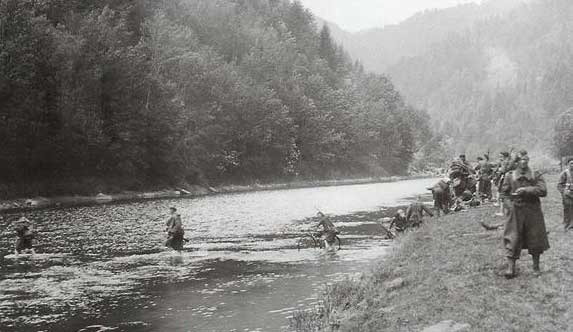 A group of soldiers cross a shallow river while others stand on the grassy riverbank surrounded by forested hills.
