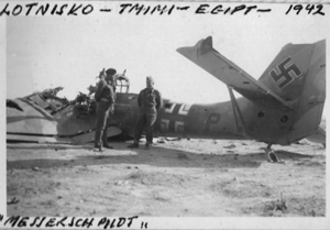 A crashed WWII German aeroplane with swastika markings lies in the desert as two soldiers stand nearby; handwritten notes label the location and year as Egypt, 1942.