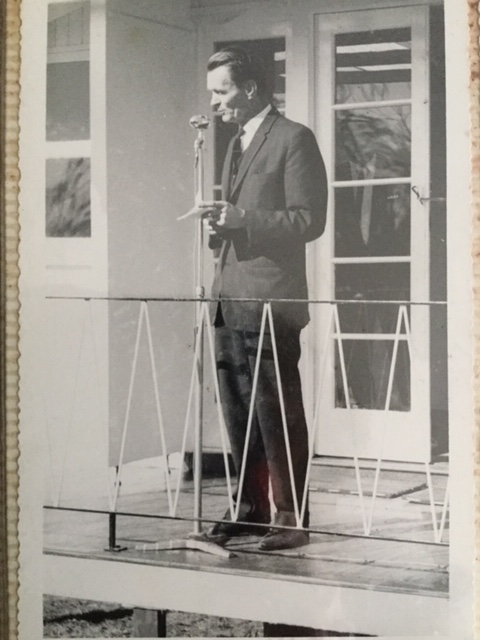 A man in a suit stands on a porch, speaking into a microphone, holding papers. There are double doors and a railing with a zigzag pattern in the background.