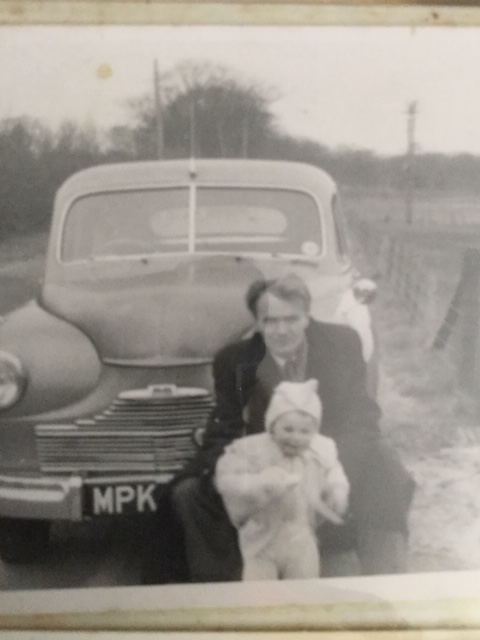 A man crouches behind a smiling baby dressed in a light outfit, posing in front of a vintage car with number plate "MPK" on a rural roadside.