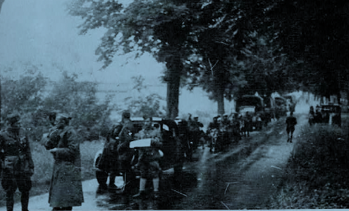 A group of soldiers with vehicles and equipment move along a tree-lined road, some standing and some walking, under overcast skies.