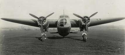 Front view of a twin-engined vintage aeroplane with a propeller on each wing, parked on a grassy field.