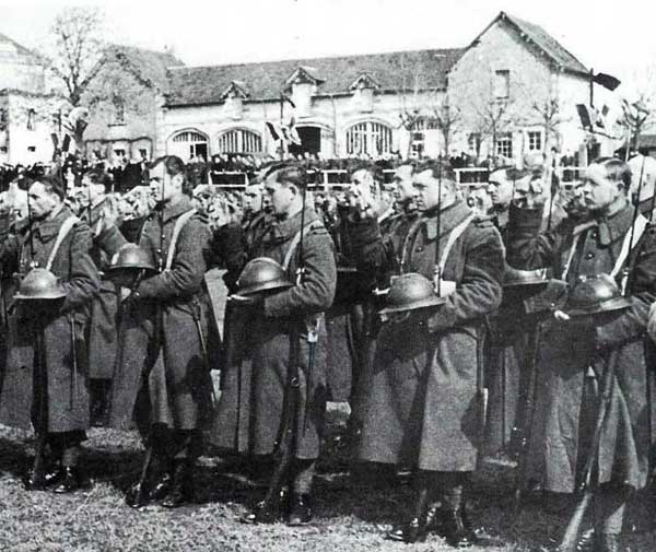 A group of soldiers in long coats and helmets stand in formation outdoors, holding rifles and helmets, with buildings and more troops in the background.