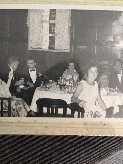Black and white photo of people in formal attire sitting at tables with food and drinks at an indoor event, with "1966" written on the image. A decorative banner hangs in the background.