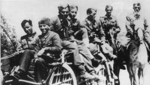 A group of soldiers wearing uniforms ride together on a motorcycle with a sidecar, whilst two others follow behind on horseback.