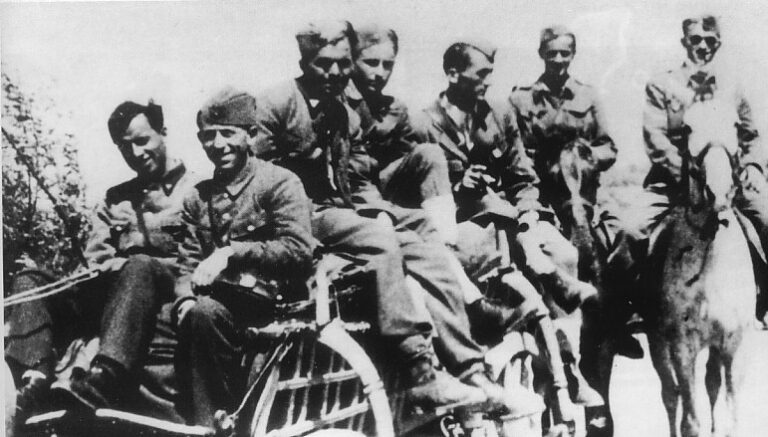 A group of soldiers wearing uniforms ride together on a motorcycle with a sidecar, whilst two others follow behind on horseback.