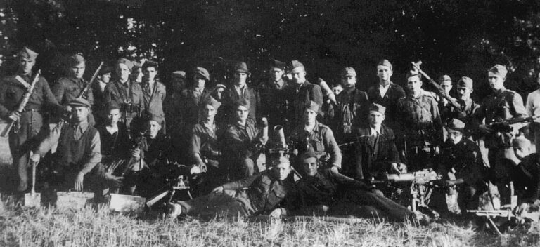 A large group of uniformed soldiers pose outdoors in black and white, some standing and some sitting or lying on the grass, holding rifles and equipment.