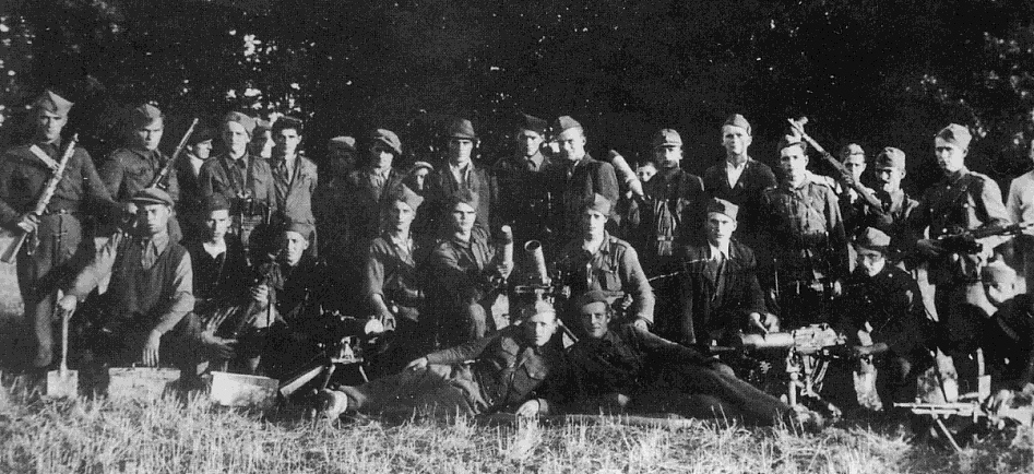 A large group of uniformed soldiers pose outdoors in black and white, some standing and some sitting or lying on the grass, holding rifles and equipment.