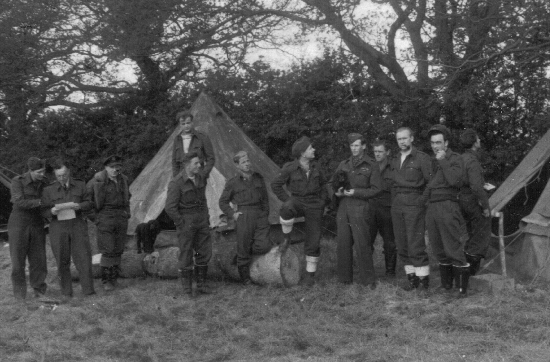 A group of soldiers in uniform stand and sit in front of tents outdoors, with trees and bushes in the background.