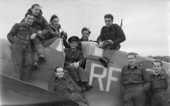 A group of eight men in military uniforms pose around and atop a vintage fighter aeroplane marked with the letters “RF.”.