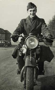A man in a military uniform sits on a motorbike, facing the camera on a tarmacked road with houses and a car in the background.