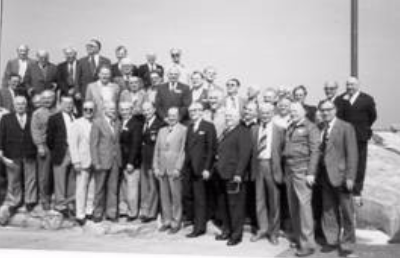 A large group of men in suits and ties pose together outdoors on a rocky surface under clear skies.