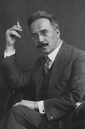 A man with glasses and a moustache sits in a chair, wearing a suit and holding a cigarette in his raised hand against a plain backdrop.