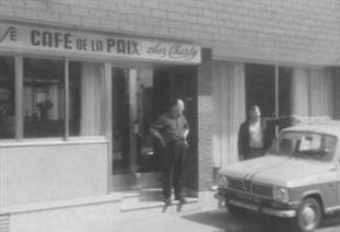 Two men stand outside a building with a sign reading "Café de la Paix" next to a parked car on the street.