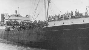 Black and white photo of a crowded ship with many people standing on deck, close to the water’s edge.