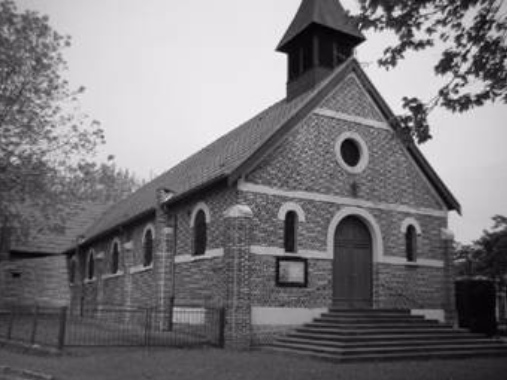 A small brick church with arched windows and a central wooden door, topped by a spire, surrounded by trees and a metal fence.