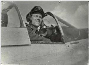 A pilot in uniform sits in the cockpit of a military aeroplane, smiling and looking outwards.