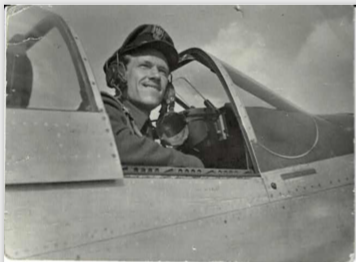 A pilot in uniform sits in the cockpit of a military aeroplane, smiling and looking outwards.