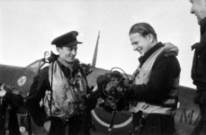 Two men in flying suits and life vests stand beside a military aeroplane, with one holding a piece of equipment and both appear to be engaged in conversation.