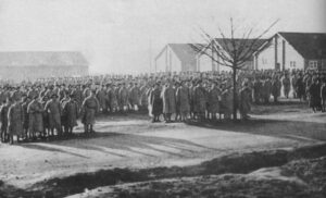 A large group of people wearing coats stands in queues outdoors near several barracks-style buildings on a clear day.