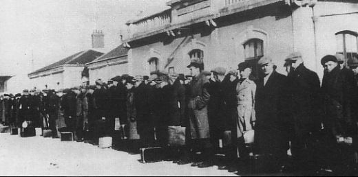 A group of men stand in a queue outdoors beside a building, many holding bags or suitcases, dressed in coats and hats, under daylight.