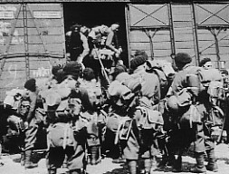 A group of soldiers with rucksacks board a railway carriage, while others stand around outside, wearing uniforms and helmets.