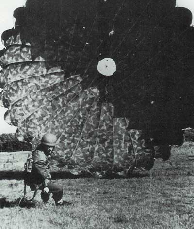A soldier in uniform kneels on grass beside a large, open parachute that has just landed.
