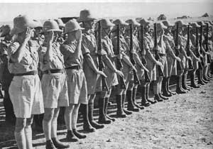 A row of soldiers in uniform and pith helmets stand to attention and salute, holding rifles, during a military formation outdoors.