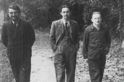 Three men in suits and jackets walk outside on a wooded path, looking towards the camera, with trees and foliage in the background.