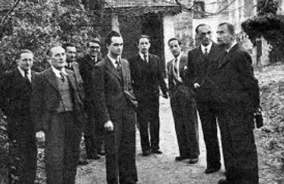 A group of men in suits stand outdoors on a path near trees and buildings, posing for a black-and-white photograph.