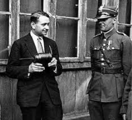 A man in a suit holding a device stands next to a uniformed officer with medals, both posed in front of a windowed building.