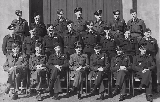 A group of uniformed military personnel pose for a formal group photo in front of a large wooden door, with some seated and others standing in rows.