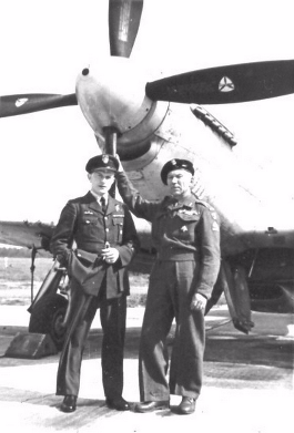 Two military men in uniform stand in front of a propeller aeroplane, one touching the propeller blade.