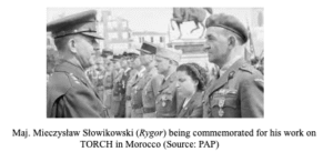 A military officer stands in uniform, receiving honours from another officer, with several people observing the ceremony outdoors.