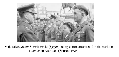A military officer stands in uniform, receiving honours from another officer, with several people observing the ceremony outdoors.