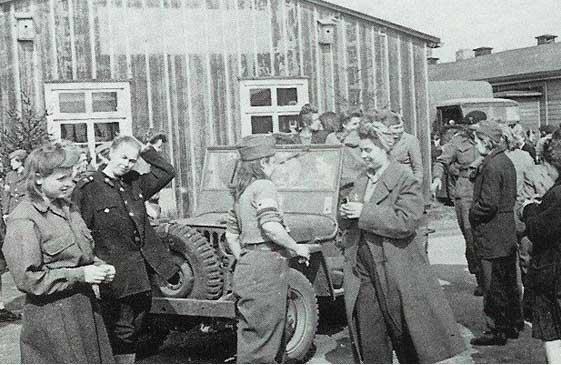 A group of people, including uniformed individuals, stand and converse near a military jeep outside a wooden building.