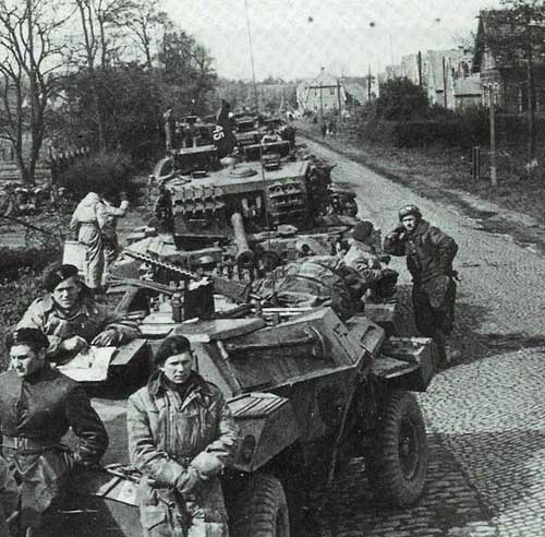 A column of military vehicles and soldiers advances along a cobbled road in a rural area; trees and buildings line both sides.