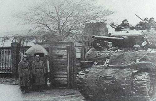 Second World War era soldiers stand beside a guard post while others ride on a tank near a gated entrance, with trees and fencing visible in the background.