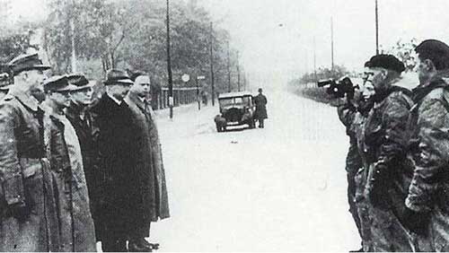 A group of men stand in a line facing armed soldiers aiming rifles on an empty street; a car is visible in the background.