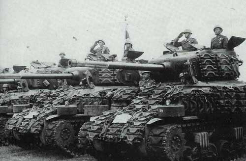 A row of soldiers in uniform are seen saluting whilst sitting atop military tanks lined up in formation.