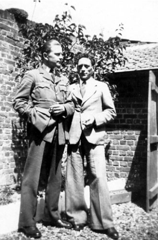 Two men in suits stand side by side in a garden area, with a brick wall and foliage in the background.