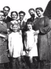 A group of five adults and two young girls pose outdoors, smiling for the camera. The adults stand behind the girls, who are both wearing light-coloured dresses.